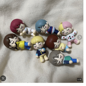 Nine colorful cartoon figurines with closed eyes and smiling faces lying on a light fabric, arranged in a loose circle.