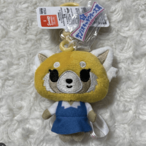 Yellow plush dog character wearing a blue dress, with white wings, lying on a fluffy white surface; a plastic tag is attached to its head.