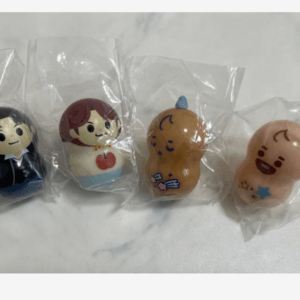 Row of four small cartoon figurines in plastic bags: a suited man, a woman with an apple on her shirt, an orange cartoon character, and a smiling bald figure with stars.
