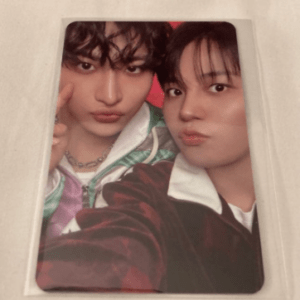 Two young people take a close-up selfie, both puckering their lips; one wears a colorful jacket and the other a dark jacket, on a light surface as a photo card.