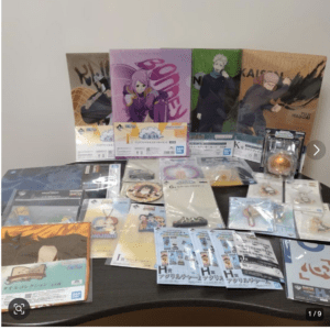 Table filled with anime merchandise: colorful Jujutsu Kaisen posters and various boxed figures arranged against a plain wall, plus smaller cards, stickers, and clear files spread across the table.