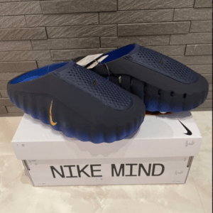 Pair of dark blue foam slide sandals with textured uppers and bright blue insoles resting on a white Nike Mind shoebox against a gray tiled wall.