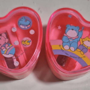 Two pink, heart-shaped plastic containers with colorful cartoon designs on the lids; the left one shows a girl on a ball and small floating shapes, the right one features a blue bear-like character and a yellow character on a rainbow with stars