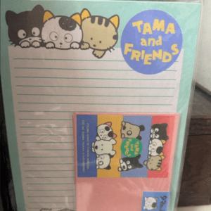 Notepad set titled 'Tama and Friends' with three cartoon cats at top, blue logo circle, and lined writing paper plus a pink card/package with cartoon characters.