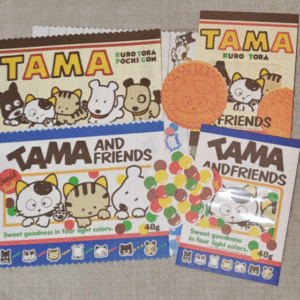 Assorted Tama and Friends snack packets with cartoon cat characters, cookies, and colorful candy balls on a fabric surface.