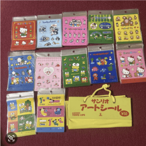 Collection of colorful character sticker sheets in clear packaging laid out on a red surface, including Hello Kitty designs, PataPataPoppy, and friends, with a yellow Japanese tote bag at the bottom right.