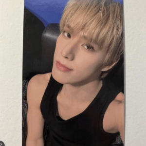 Close-up selfie of a person with short blond hair wearing a black sleeveless shirt, sitting in a chair with a blue background behind them.