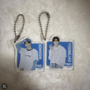 Two acrylic keychains with ball chains, each featuring a photo of a young man in white clothing on a blue background; left keychain reads 'DOHOON', right keychain reads 'YOUNGJAE' with the person waving on the right.