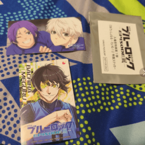 Three anime cards laid on a blue patterned bedsheet: purple- and white-haired characters on the top card, a black-haired hero on the bottom card, and a gray sleeve with Japanese text on the right.