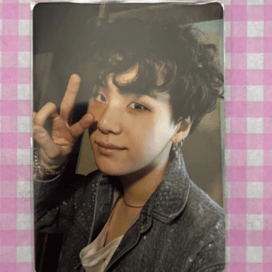 Close-up portrait of a young man with dark curls making a peace sign, wearing a denim jacket and earring, against a soft backdrop.