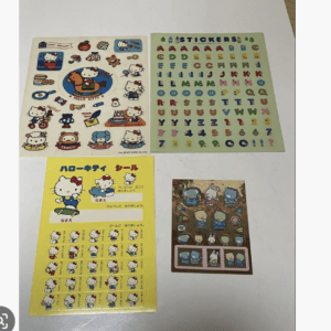 Four sticker sheets laid out on a light surface: Hello Kitty characters, colorful alphabet sheet, and a yellow instruction sheet in the corner.