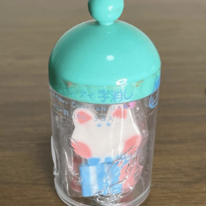 Clear plastic capsule toy with a teal cap and top knob, housing a small red and blue figure inside.