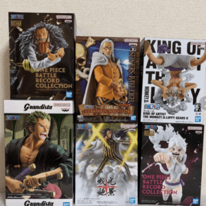 Boxed anime figures from One Piece, Battle Record Collection and Grandista lineup on a beige fabric backdrop edge of a shelf/board grip area.
