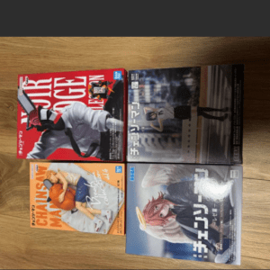 Four manga magazines laid on a wooden floor: two stacked on the left (red and orange covers) and two on the right (gray and blue covers) featuring anime characters.
