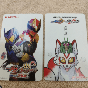 Two illustrated cards on a beige surface: left card features Ultraman heroes, right card shows a white fox-like creature with red markings and yellow eyes