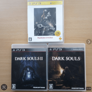 Three PS3 game cases on a wooden table: Demon's Souls in a white case above, with Dark Souls and Dark Souls II in black cases below (FromSoftware).