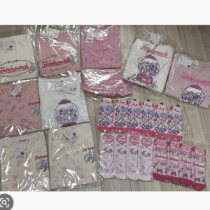 Assorted pink and white children's clothes in clear packaging, laid out on a wooden floor with colorful gumball machine graphics.