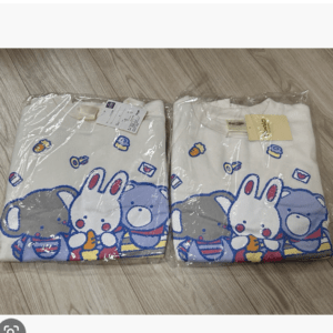 Two white t-shirts with cartoon rabbits and a bear in plastic packaging on a light wood floor surface.