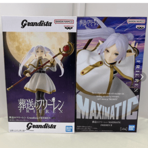 Two boxed anime figures side by side: left box shows a white outfit figure holding a staff with a large moon background, labeled Grandista Frieren by Bandai Namco; right box features a white-haired elf figure in a dynamic pose against a purple gradient, labeled MAXIMATIC Frieren II by Bandai Namco.
