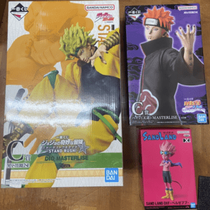Three boxed anime figure sets: a large Dio Brando/Masterile box on the left with a yellow-and-green character, Bandai Namco branding visible; a purple Naruto Masterline figure box on the top right; a pink Sand Land figure box at the bottom right showing a pink-haired character with goggles.