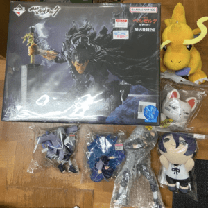 Large boxed action figure of a battle-worn, armored warrior wielding a massive sword on a wooden floor, with small anime figures in bags and a yellow dragon plush nearby.