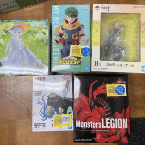 Assorted boxed anime figures laid out on a wooden floor, including a My Hero Academia Deku figure box and a red Dragon Monsters Legion box.