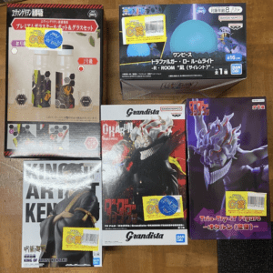 Collection of Japanese anime figures in boxed packaging on a wooden surface, including Grandista and King of Artist Kenshiro boxes by Bandai Namco/Good figures sets