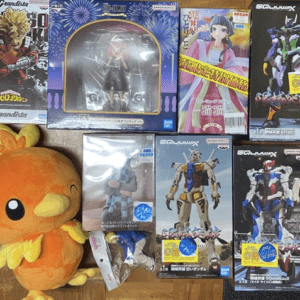 Orange plush chick sits beside a collection of boxed anime and Gundam action figures on a wooden floor.