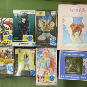 Assorted boxed anime figurines arranged on a green surface, including a pink-winged angel figurine box on the right and several tall character boxes in the center and left.
