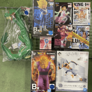 Stack of boxed anime action figures on a display rack, featuring Dragon Ball Z characters (including a Super Saiyan) and One Piece figures, plus a large green dragon toy in plastic wrapping on the left.