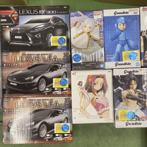 Row of boxed scale models and anime figures on a green tabletop, including Lexus cars and Grandista figures.
