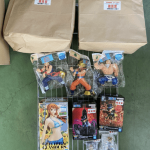 Wire display rack on a green floor with two large cardboard boxes on top; three plastic-wrapped action figures and several boxed anime figures on lower shelves.