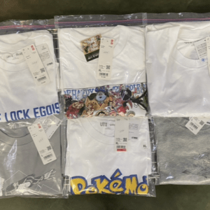 Six white graphic T-shirts in clear plastic bags hanging on a metal grid rack, each with colorful designs visible (anime, Pokémon, text).