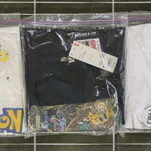 Three folded T-shirts in plastic bags on a metal rack: left white with a large Pokémon logo, middle black shirt, right white shirt with a purple feather-like graphic and Japanese text, price tags attached.  Articulos Japan (Post Box Japan)
