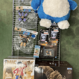 Metal display rack with a large blue-and-white Snorlax plush at the top right, plus hanging anime figure plush and figures in plastic bags along the left side, on a green floor; several boxed collectibles sit at the bottom.