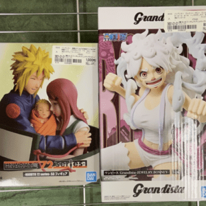 Two boxed anime figures on a metal display rack; left box shows a blond spiky-haired character cradling a baby with a pink-haired figure nearby, right box features a white-haired character in a white outfit with an energetic pose and fists. Price tags are visible on both boxes.