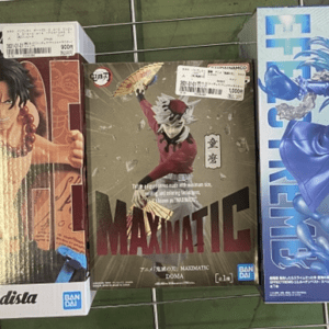 Three boxed anime figure sets on a green grid shelf: left is Grandista Grandista box with a shirtless muscular character, center Maximatic figure, right blue-haired dynamic character in blue armor/robe.