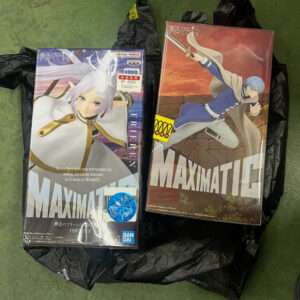 Two boxed anime figures on a black bag on a green floor; one white-haired character in white/gold outfit, the other blue-haired in a blue outfit labeled MAXIMATIC by Bandai Namco.