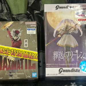 Two boxed anime figures sit on a black plastic bag on a green surface: the left box shows a red-hooded character with bold MAXIMATIC text, the right box features a white-dressed female figure with a staff under a moonlit backdrop and Grandista branding.