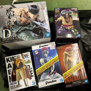 Collection of anime figure boxes on a black bag: large Zoro sword stance figure and several One Piece Grandista boxes visible, yellow and pink price stickers present.