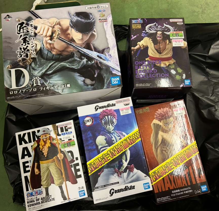 Img_0232 - Articulos Japan (post Box Japan) Collection of anime figure boxes on a black bag: large Zoro sword stance figure and several One Piece Grandista boxes visible, yellow and pink price stickers present. Articulos Japan (Post Box Japan)