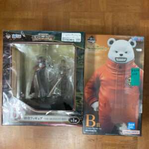 Two boxed anime figures on a wooden floor: left is a detailed character figurine in a display box, right is a bear-headed figure in an orange suit in its box.