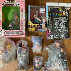 Assorted anime action figures and boxes on a wooden floor, including a pink One Piece box with a green figure inside and wrapped figures nearby.