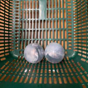 Two translucent plastic balls resting side by side in a green perforated basket with rectangular openings for ventilation.