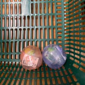Two translucent toy capsules in a green shopping basket, one pinkish and one purple, each with wrapped contents inside.