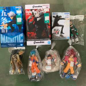 Boxes of anime figures (Maximatic and Grandista) with loose figures in plastic bags laid on a green floor, price tags visible on packaging.