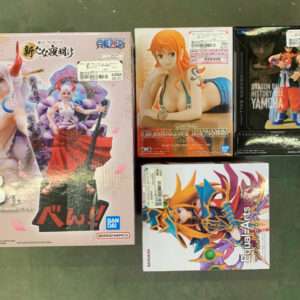 Box of four anime figurines on a green floor: large left box with pink-haired characters, center box with a woman in a swimsuit, top-right Dragon Ball Yamcha box, bottom-right colorful armored figure box.