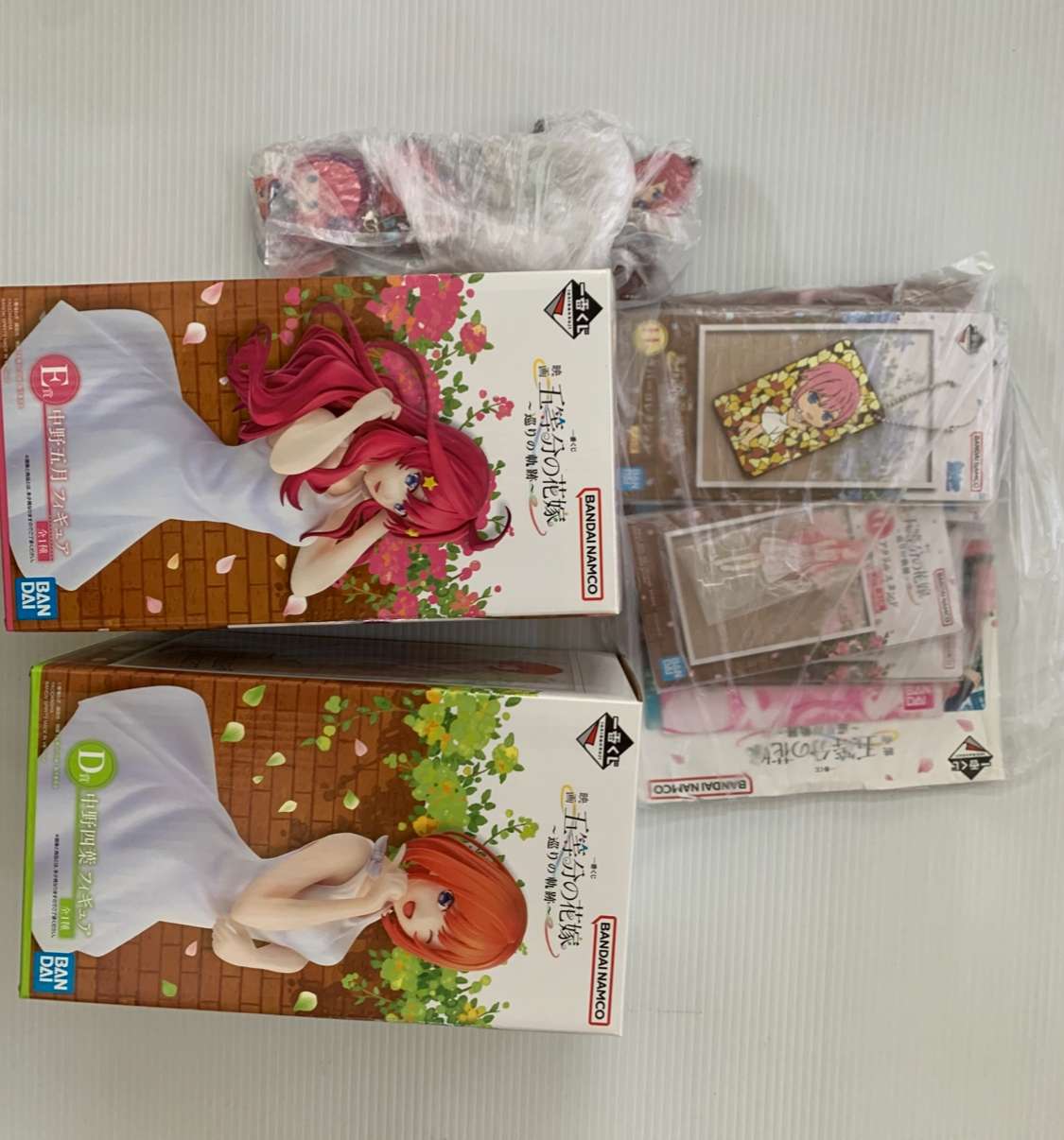 Img_2074 - Articulos Japan (post Box Japan) Two Bandai Namco anime figure boxes showing a pink-haired girl and an orange-haired girl in white dresses amid a brick wall and flowers; other figure boxes and accessories are wrapped nearby. Articulos Japan (Post Box Japan)