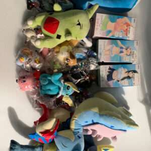 Assorted plush toys and a blue U-shaped neck pillow lying on a white surface, with various character plushies and small boxed items nearby.