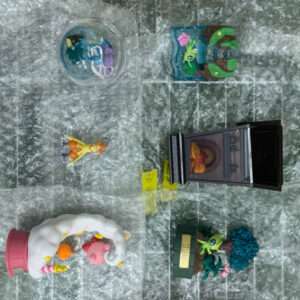Assorted colorful plastic toy figures and small diorama pieces spread on bubble wrap, including a person in orange, a dragon-like figure, and a display stand area.